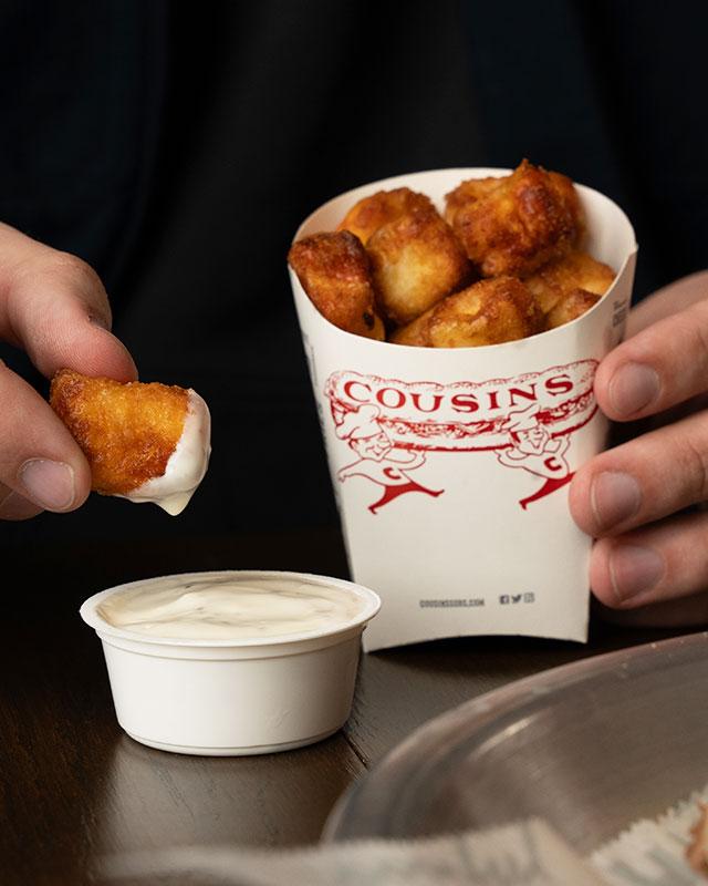 Hand holding a cheese curd dipping it in sauce with Cousins Subs container in the other hand.